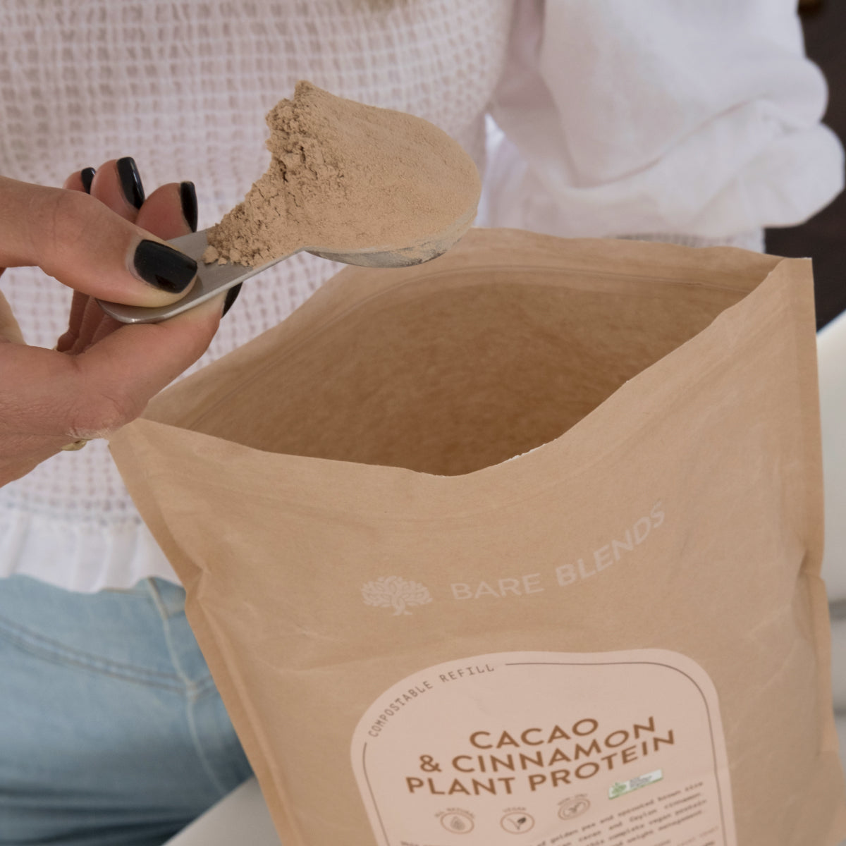 Scoop of Cacao and Cinnamon Plant Protein powder held above an open Bare Blends pouch