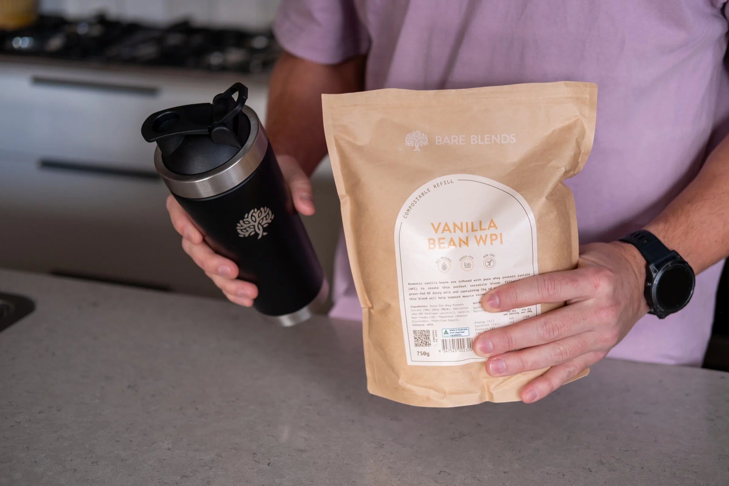Person holding Bare Blends Vanilla Bean WPI and a black Bare Blends shaker in a kitchen